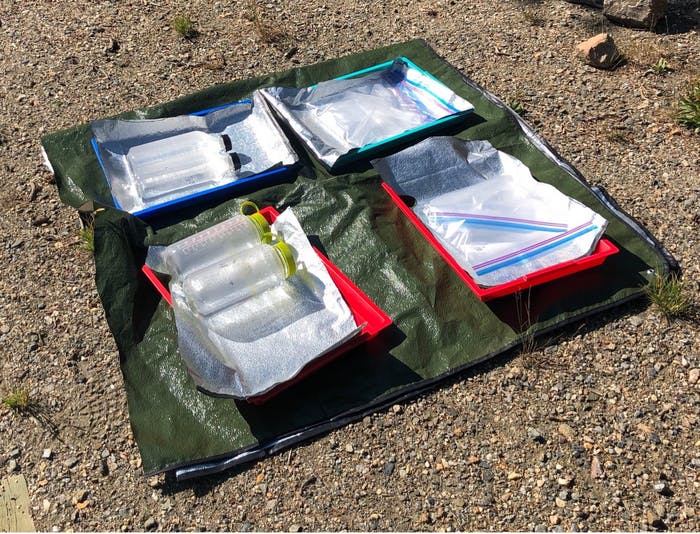 An experimental setup with Nalgene bottles, plastic water bottles, and a plastic bag placed on a reflective tarp exposed to the sunlight during a lower-altitude trial.