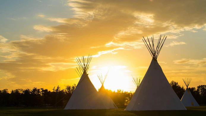 An array of indigenous tee-pees on the prairie grass set up for a cultural event at sunset.